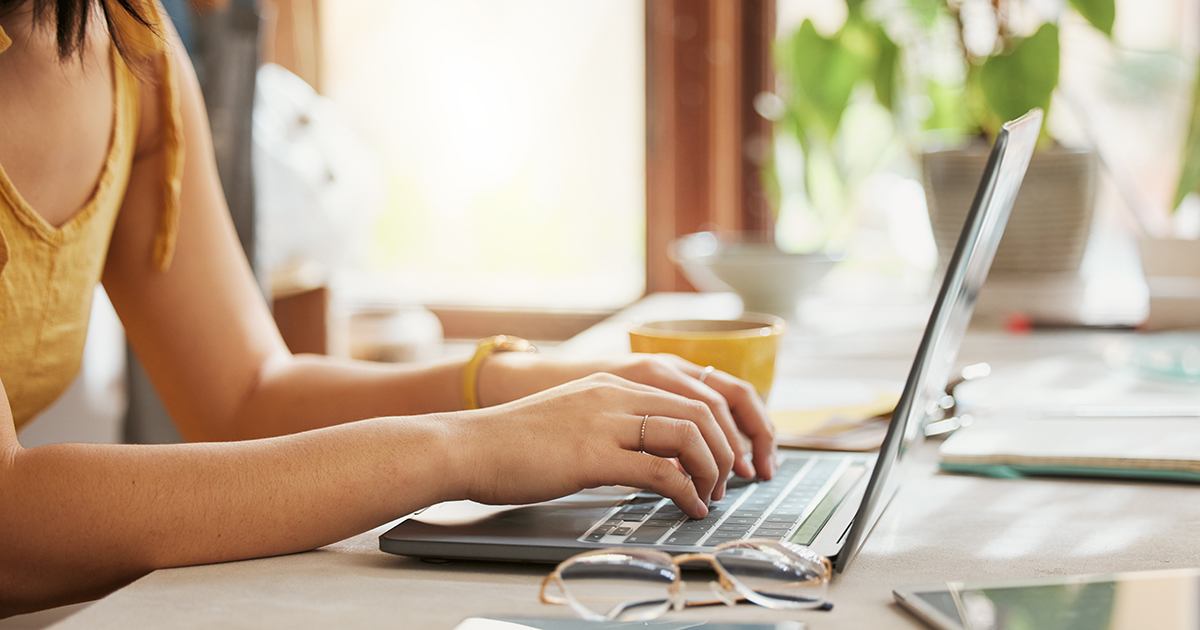 Person typing on a laptop at a sunlit table with a mug, papers, and plants in the background.